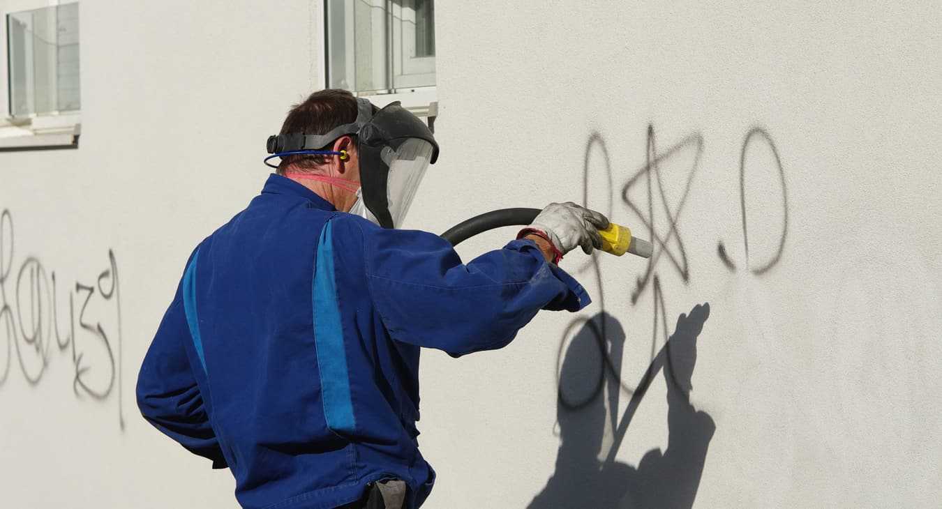 Arbeiter in blauem Schutzanzug entfernt mit Hochdruckverfahren Grafitti auf Hauswand.